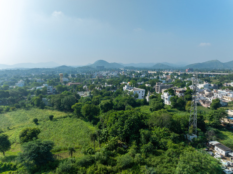 Aerial view of the cityscape of Udaipur Rajasthan India featuring lush green trees residential buildings and a tall communication tower under a clear blue sky. Aerial view of the cityscape of Udaipur Rajasthan India featuring lush green trees residential buildings and a tall communication tower under a clear blue sky.