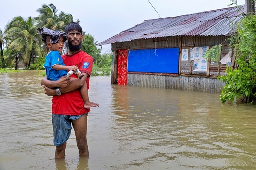 Banshkhali Upazila Chittagong Division Bangladesh 10 July 2026 View of a man carrying a crying child through floodwaters in front of a small house in a rural area. Banshkhali Upazila Chittagong Division Bangladesh 10 July 2026 View of a man carrying a crying child through floodwaters in front of a small house in a rural area.