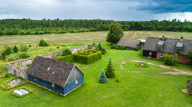 Aerial view of a traditional rural farmstead featuring wooden houses with shingle roofs stone wall ruins and a lush green lawn surrounded by fields and forest in Latvia. Aerial view of a traditional rural farmstead featuring wooden houses with shingle roofs stone wall ruins and a lush green lawn surrounded by fields and forest in Latvia.