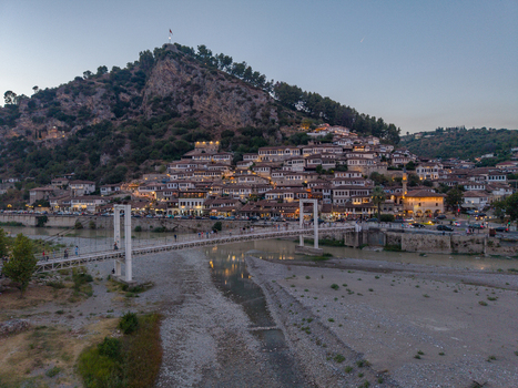 Aerial view of the historic hillside houses of Berat featuring the Osum River and a pedestrian suspension bridge under a soft twilight sky in Berat Berat County Albania.