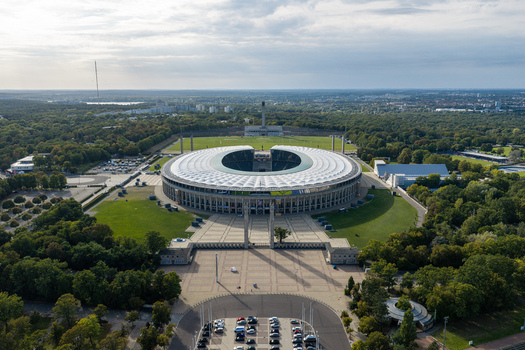 Berlin Germany 26 September 2026 Aerial view of the Olympiastadion Berlin showing its iconic circular roof structure surrounding green parkland and the expansive plaza under a bright sky.