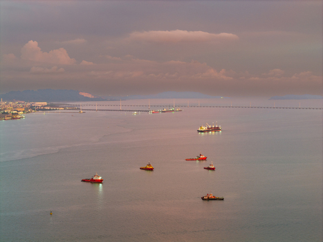 Aerial view of cargo ships and tugboats navigating the calm waters of the Penang Strait with the Penang Bridge in the distance under a soft sunset sky in George Town Penang Malaysia.