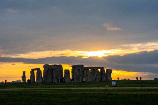 View of Stonehenge at sunset with silhouetted stones against a vibrant orange sky and people walking in the surrounding field in Salisbury England United Kingdom.