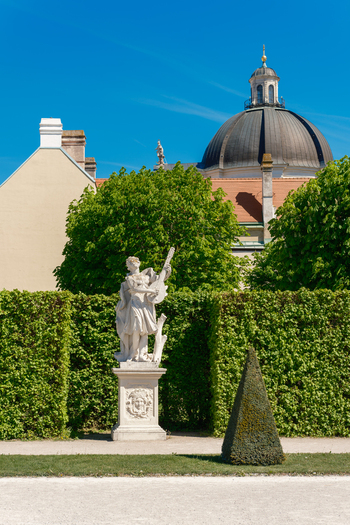 View of the Belvedere Palace gardens featuring a baroque stone statue of a musician in front of manicured hedges and a historic dome under a clear blue sky in Vienna Vienna Austria. View of the Belvedere Palace gardens featuring a baroque stone statue of a musician in front of manicured hedges and a historic dome under a clear blue sky in Vienna Vienna Austria.