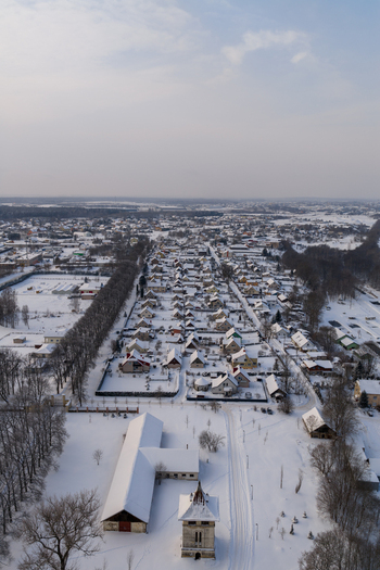 Aerial view of a snow covered village featuring residential houses bare trees and a historic tower building under a pale winter sky Lithuania. Aerial view of a snow covered village featuring residential houses bare trees and a historic tower building under a pale winter sky Lithuania.