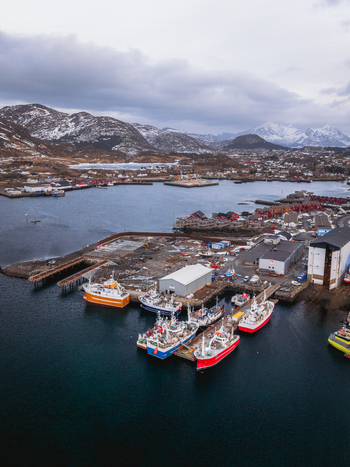Aerial view of boats clustered in the harbor against a backdrop of snow dusted mountains and a cloudy sky Ballstad Nordland Norway. Aerial view of boats clustered in the harbor against a backdrop of snow dusted mountains and a cloudy sky Ballstad Nordland Norway.