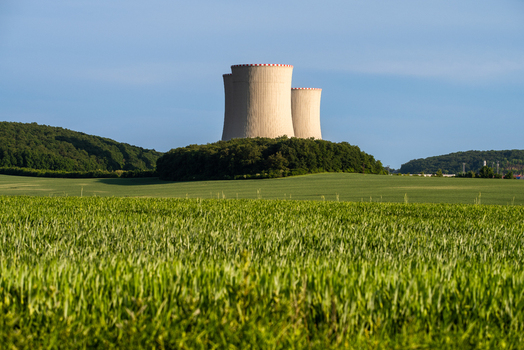 View of the cooling towers of a nuclear power plant rising above a lush green field and surrounding trees under a clear blue sky in Czechia. View of the cooling towers of a nuclear power plant rising above a lush green field and surrounding trees under a clear blue sky in Czechia.