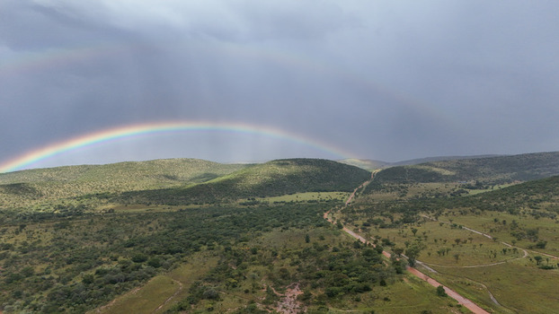 Aerial view of a vibrant rainbow stretching across rolling green hills and a dirt road under a stormy sky in Waterberg District Municipality Limpopo South Africa. Aerial view of a vibrant rainbow stretching across rolling green hills and a dirt road under a stormy sky in Waterberg District Municipality Limpopo South Africa.