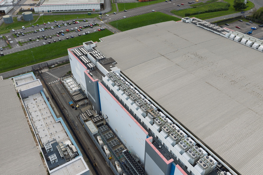 Aerial view of a large data center facility featuring extensive rooftop cooling units adjacent parking areas and surrounding industrial infrastructure in the United Kingdom.