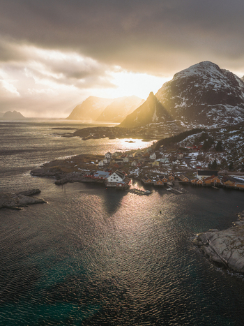 Aerial view of golden sunlight piercing through dramatic clouds illuminating the rugged coastline and quaint village nestled below snow dusted mountains Tind Nordland Norway.