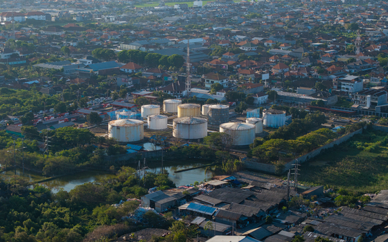 Aerial view of industrial fuel storage tanks surrounded by urban residential buildings lush trees and a small pond in Kecamatan Denpasar Selatan Bali Indonesia.