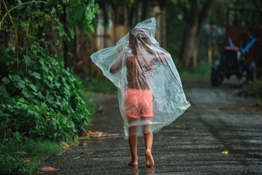 Bogura Rajshahi Division Bangladesh 07 July 2019 View of a child walking barefoot on a wet road during a rain shower wearing a makeshift plastic poncho.