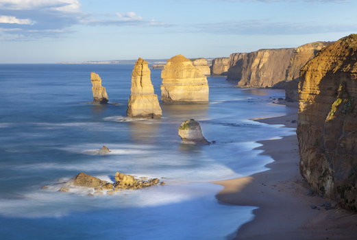 View of the Twelve Apostles limestone stacks standing in the ocean along the rugged coastline with steep cliffs and a sandy beach under a clear sky in Australia. View of the Twelve Apostles limestone stacks standing in the ocean along the rugged coastline with steep cliffs and a sandy beach under a clear sky in Australia.
