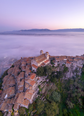 Aerial view of the historic town of Artena perched on a rocky cliffside with terracotta rooftops and a church rising above a thick layer of morning mist in Artena Lazio Italy.