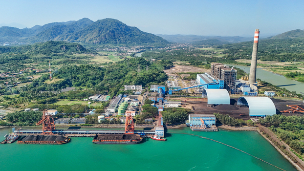 Aerial view of a coal fired power plant with a tall chimney coal storage domes and a river terminal with barges in Indonesia.