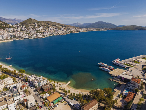 Aerial view of the coastal city of Saranda featuring a busy harbor with ferries a sandy beach and urban architecture set against rolling mountains in Saranda Vlore County Albania. Aerial view of the coastal city of Saranda featuring a busy harbor with ferries a sandy beach and urban architecture set against rolling mountains in Saranda Vlore County Albania.