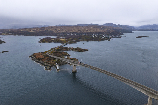 Aerial view of the Skye Bridge spanning across the sea with the Eilean Ban lighthouse and rugged hills under an overcast sky Isle of Skye Scotland United Kingdom.