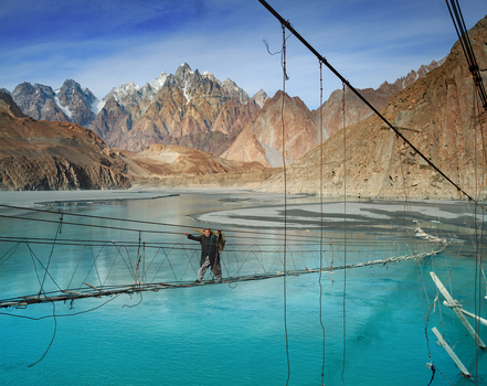 Upper Hunza Gilgit Baltistan Pakistan 08 February 2013 View of a man crossing the Hussaini Suspension Bridge over turquoise river waters with rugged mountain peaks.