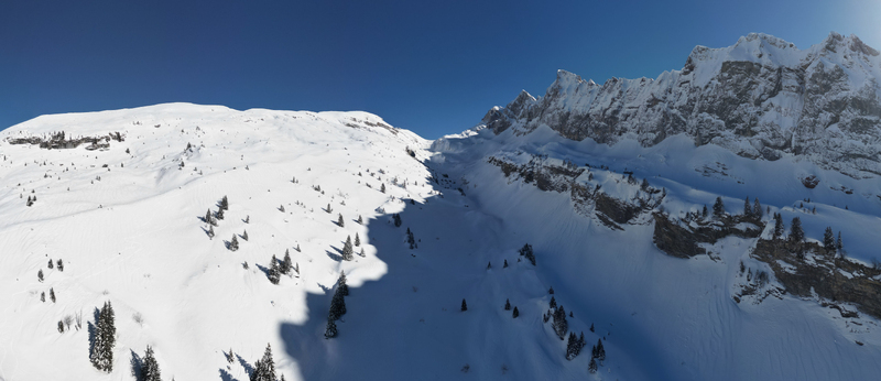 Aerial view of snow covered mountain ranges with a scattering of dark green trees under a clear blue sky Samoëns Auvergne Rhône Alpes France. Aerial view of snow covered mountain ranges with a scattering of dark green trees under a clear blue sky Samoëns Auvergne Rhône Alpes France.