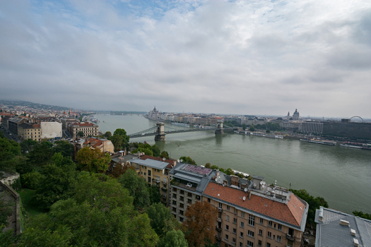 View of the Szechenyi Chain Bridge spanning the Danube River with historic city architecture and rooftops under a cloudy sky in Budapest Hungary.