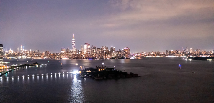 New York New York United States 11 August 2021 Aerial view of the Manhattan skyline at night with Ellis Island in the foreground and the Hudson River reflecting city lights.