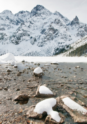 View of Morskie Oko lake surrounded by snow covered Tatra Mountains with rocky peaks and snow capped stones in the foreground Tatra County Lesser Poland Poland. View of Morskie Oko lake surrounded by snow covered Tatra Mountains with rocky peaks and snow capped stones in the foreground Tatra County Lesser Poland Poland.