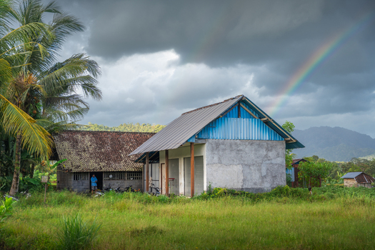 View of traditional rural houses surrounded by palm trees and lush greenery with a vibrant rainbow against a dark cloudy sky Sidemen Bali Indonesia. View of traditional rural houses surrounded by palm trees and lush greenery with a vibrant rainbow against a dark cloudy sky Sidemen Bali Indonesia.