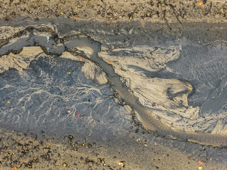 Aerial view of a textured riverbed with dark silt and light sand patterns scattered autumn leaves and natural erosion formations in Latvia.