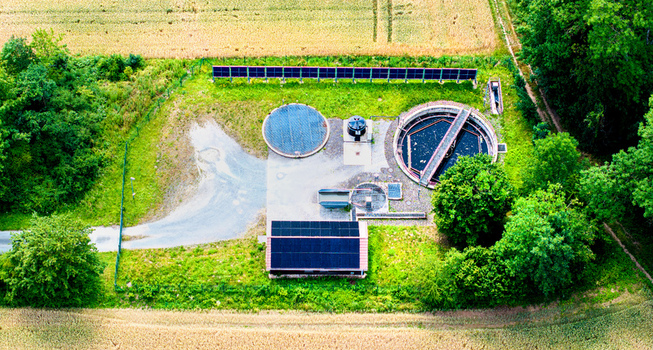 Aerial view of a small wastewater treatment plant featuring circular sedimentation tanks and solar panels on the roof situated amidst agricultural fields and trees in Germany.