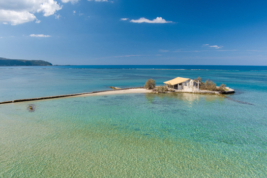 Aerial view of a small chapel on a tiny islet connected by a stone path to the shore surrounded by clear turquoise water in Lefkada Greece.