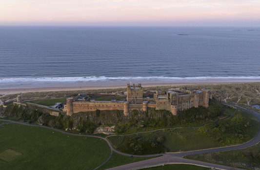 Aerial view of Bamburgh Castle standing on a rocky plateau overlooking the sandy beach and the North Sea under a soft sunset sky in Bamburgh England United Kingdom. Aerial view of Bamburgh Castle standing on a rocky plateau overlooking the sandy beach and the North Sea under a soft sunset sky in Bamburgh England United Kingdom.