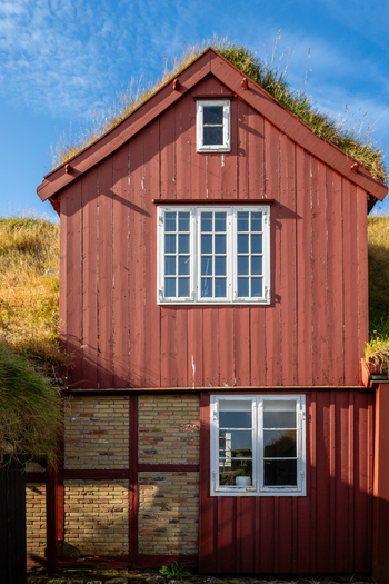 View of a traditional red wooden house with a green turf roof and brick lower level under a clear blue sky in the Faroe Islands. View of a traditional red wooden house with a green turf roof and brick lower level under a clear blue sky in the Faroe Islands.