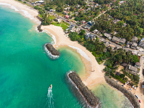 Aerial view of the tropical coastline featuring turquoise ocean water sandy beaches and rock breakwaters near a lush palm fringed road in Peraliya Southern Province Sri Lanka. Aerial view of the tropical coastline featuring turquoise ocean water sandy beaches and rock breakwaters near a lush palm fringed road in Peraliya Southern Province Sri Lanka.