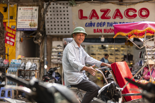 Hanoi Vietnam 11 November 2025 View of a smiling man in a green pith helmet sitting on a traditional cyclo in a busy street with shop signs and motorcycles in the background.