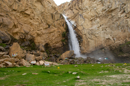 View of Khamosh Waterfall cascading down steep rocky cliffs into a green meadow with scattered boulders in Skardu Gilgit Pakistan.