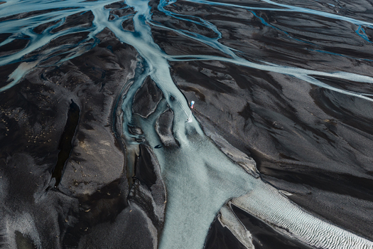 Aerial view of a kite surfer gliding over the turquoise glacial waters of a braided river winding through the dark volcanic sand landscape of Skaftarhreppur Iceland.