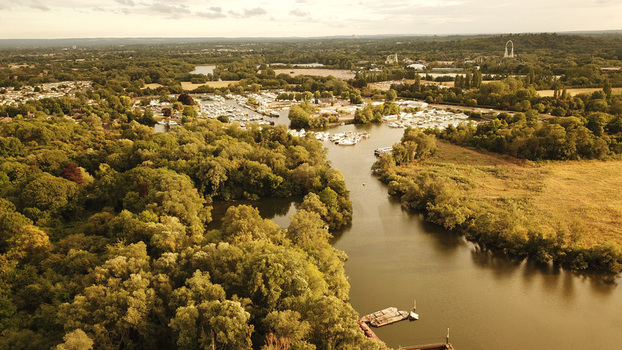 Aerial view of the Thames River winding through lush green trees and vegetation featuring a marina with moored boats and a distant landscape in Thames United Kingdom. Aerial view of the Thames River winding through lush green trees and vegetation featuring a marina with moored boats and a distant landscape in Thames United Kingdom.