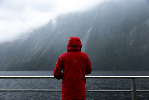 View of a person in a bright red rain jacket gazing out at the tranquil dark waters against a backdrop of majestic mist shrouded mountains Milford Sound Southland Region New Zealand.