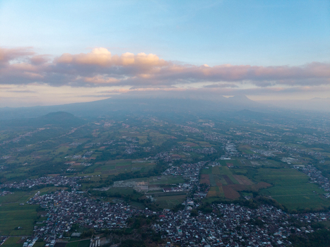 Aerial view of Malang city nestled among lush green fields and agricultural lands with a distant mountain peak under a soft sky in Malang East Java Indonesia.