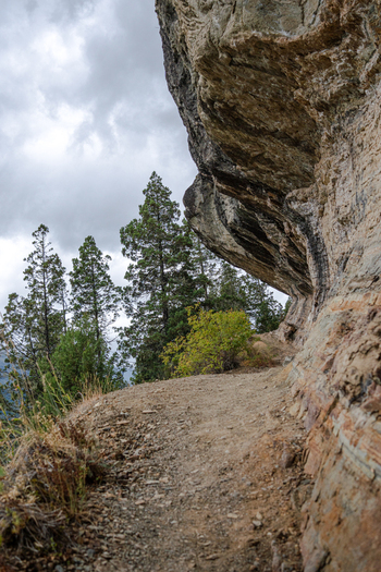 View of the hiking trail beneath the rocky overhang of Indian Head surrounded by evergreen trees under a cloudy sky in Bariloche Department Rio Negro Province Argentina. View of the hiking trail beneath the rocky overhang of Indian Head surrounded by evergreen trees under a cloudy sky in Bariloche Department Rio Negro Province Argentina.