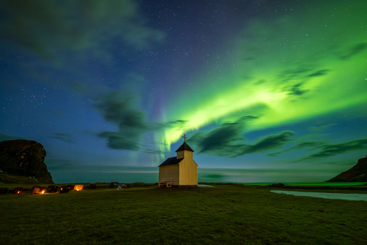 View of a small wooden church illuminated under a vibrant green aurora borealis in a grassy landscape at night in Leknes Lofoten Norway.