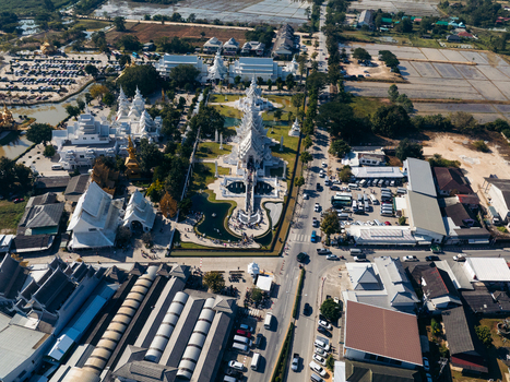Aerial view of the intricate white architecture of Wat Rong Khun temple complex surrounded by lush gardens and urban structures in Pa O Don Chai Chiang Rai Thailand. Aerial view of the intricate white architecture of Wat Rong Khun temple complex surrounded by lush gardens and urban structures in Pa O Don Chai Chiang Rai Thailand.