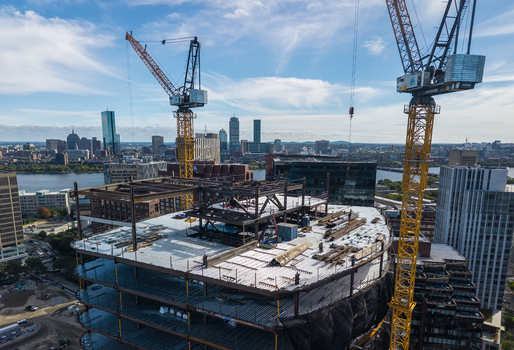 Aerial view of the Kendall Square construction site featuring steel framework and tall yellow cranes under a bright blue sky in Cambridge Massachusetts United States. Aerial view of the Kendall Square construction site featuring steel framework and tall yellow cranes under a bright blue sky in Cambridge Massachusetts United States.
