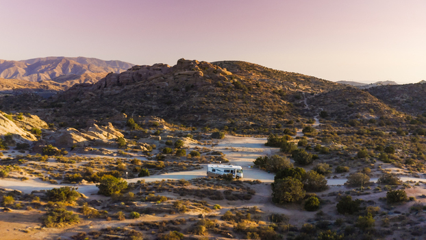 Aerial view of a recreational vehicle parked in a desert landscape with rocky hills sparse vegetation and a dirt path under a clear sky at golden hour in the United States. Aerial view of a recreational vehicle parked in a desert landscape with rocky hills sparse vegetation and a dirt path under a clear sky at golden hour in the United States.