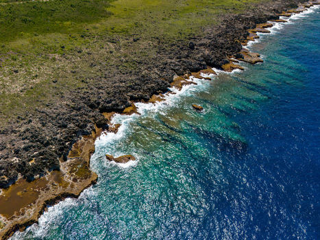 Aerial view of Chigit Beach featuring rugged volcanic rocks turquoise shallow waters and deep blue ocean waves in Northern Tinian Tinian Northern Mariana Islands. Aerial view of Chigit Beach featuring rugged volcanic rocks turquoise shallow waters and deep blue ocean waves in Northern Tinian Tinian Northern Mariana Islands.