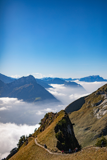 Aerial view of the Swiss Alps mountain ridge with a hiking trail people walking along the path and clouds filling the valley below in Airolo Ticino Switzerland. Aerial view of the Swiss Alps mountain ridge with a hiking trail people walking along the path and clouds filling the valley below in Airolo Ticino Switzerland.