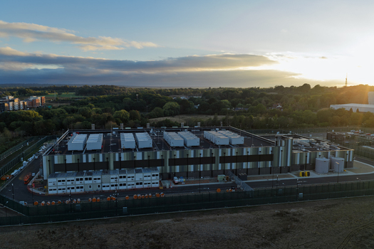 Aerial view of a modern data center facility with rooftop cooling units and surrounding security fencing set against a sunset sky in the United Kingdom. Aerial view of a modern data center facility with rooftop cooling units and surrounding security fencing set against a sunset sky in the United Kingdom.