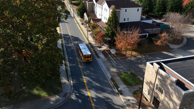 Aerial view of a yellow school bus driving along a quiet road surrounded by autumn trees and suburban buildings in Lynchburg Virginia United States. Aerial view of a yellow school bus driving along a quiet road surrounded by autumn trees and suburban buildings in Lynchburg Virginia United States.