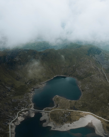 Aerial view of Snowdon mountain landscape featuring a dark glacial lake surrounded by rugged rocky slopes and winding paths under a cloudy sky in Caernarfon Wales United Kingdom.