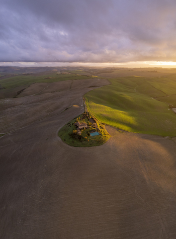 Aerial view of a farmhouse with a swimming pool and cypress trees surrounded by rolling hills in the golden hour light in Asciano Tuscany Italy.